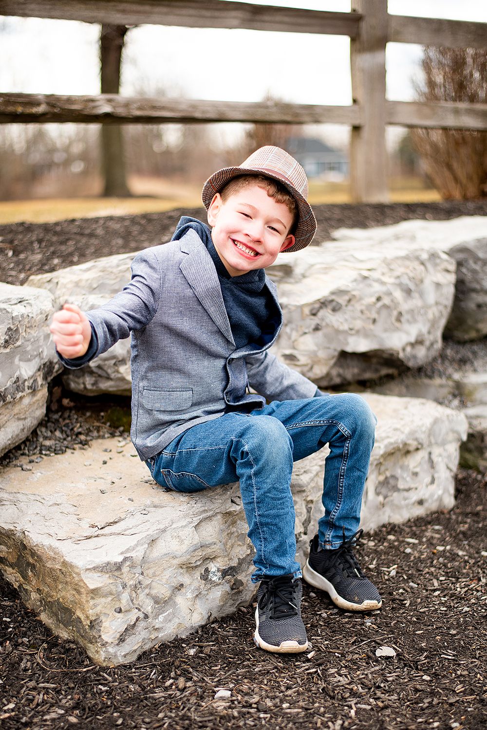 A young child in a gray blazer and fedora hat sits casually on large rocks while smiling outdoors.
