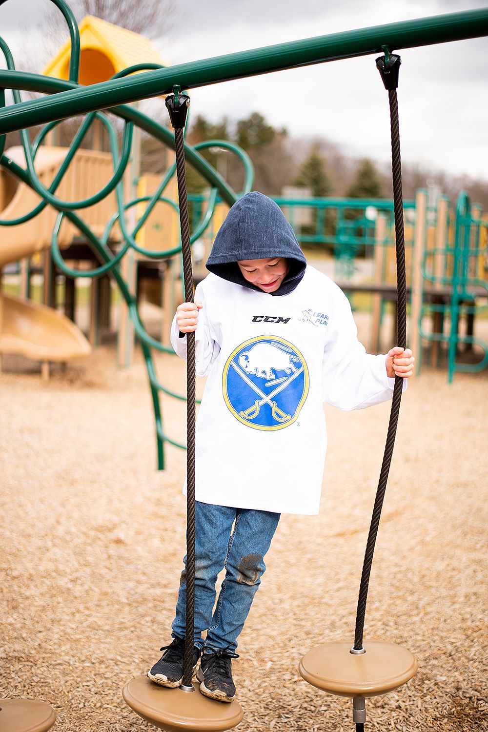 Young hockey fan in Buffalo Sabres shirt plays on playground equipment at a park on a cloudy day.
