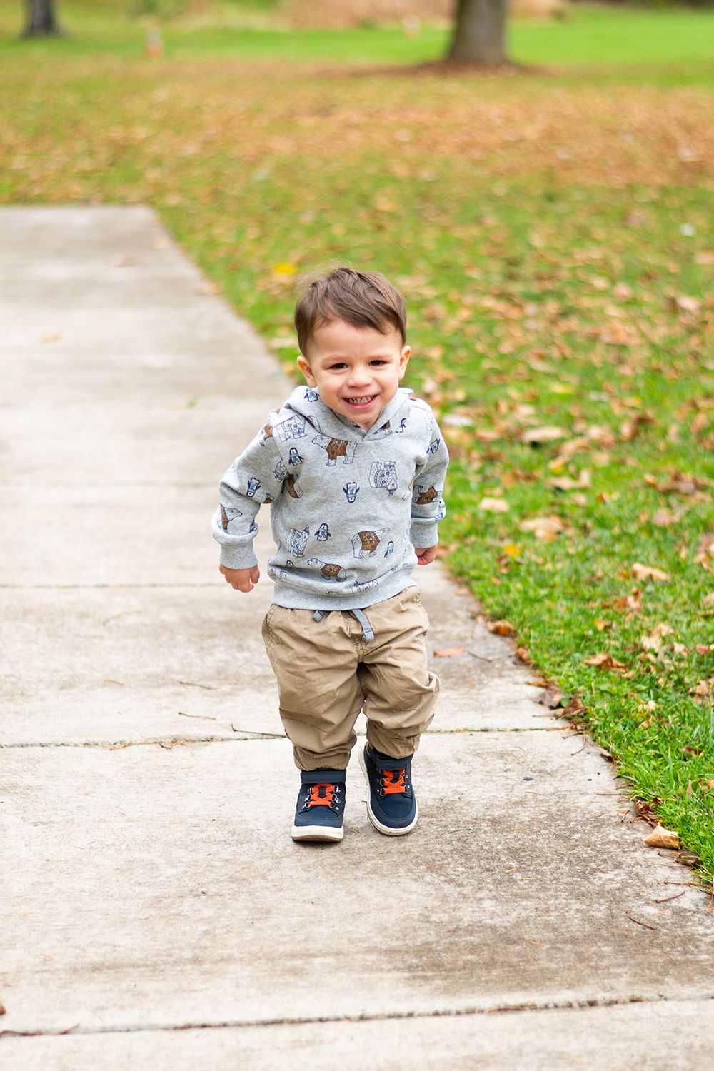 A toddler in khaki pants and a printed hoodie runs joyfully on a concrete path surrounded by autumn leaves.