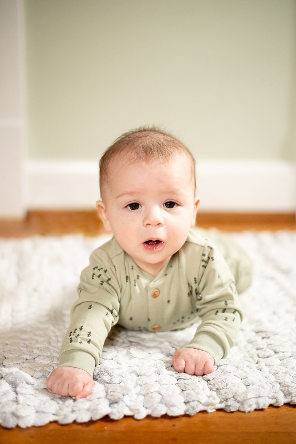 Baby in sage green outfit lies on textured white rug while doing tummy time on hardwood floor.