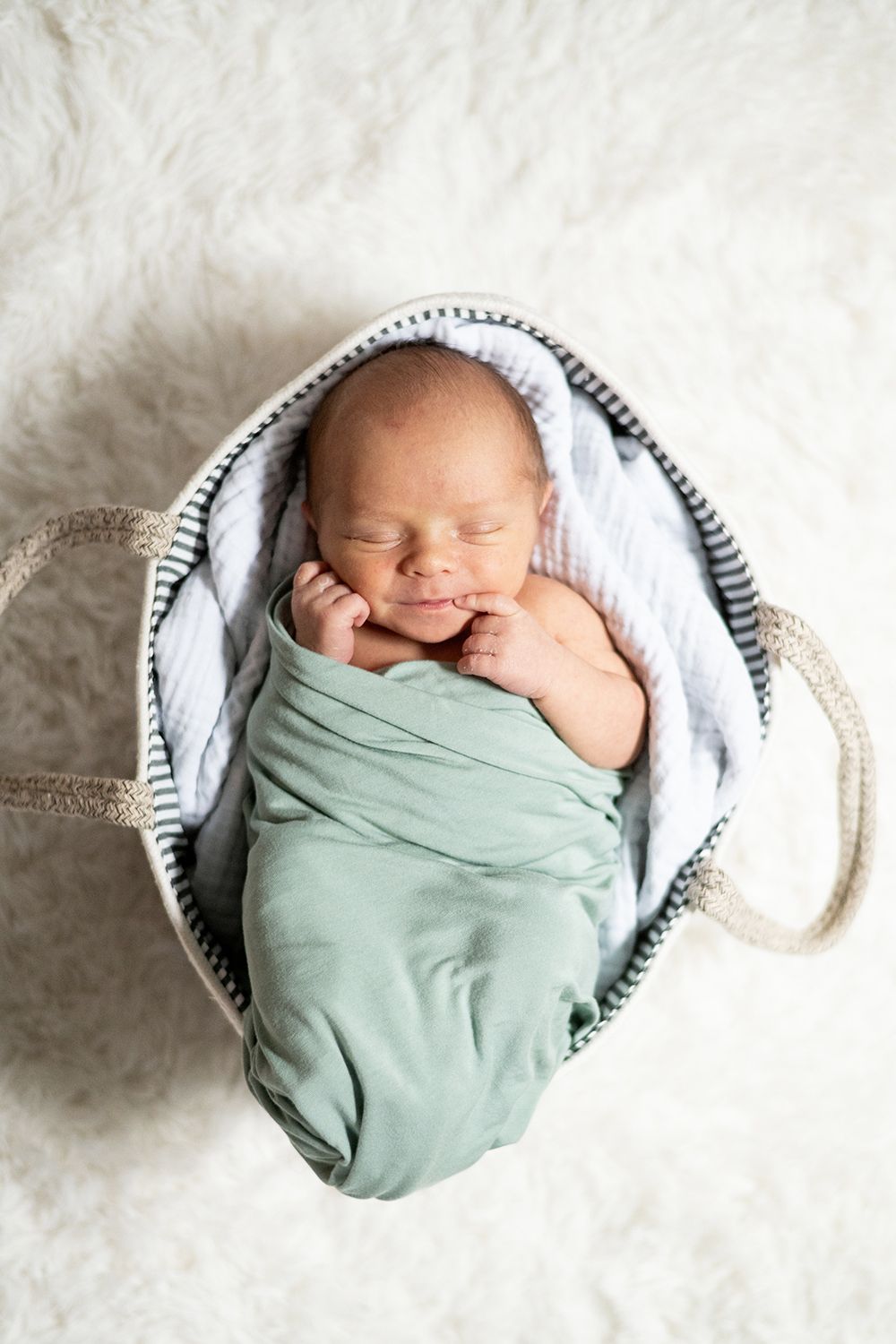 Sleeping newborn swaddled in mint green wrap lies peacefully in striped basket on white fur blanket.