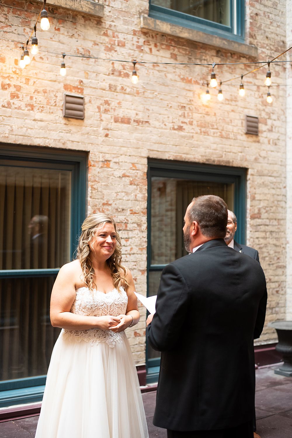 A couple shares their first look moment in a rustic brick venue with string lights overhead.