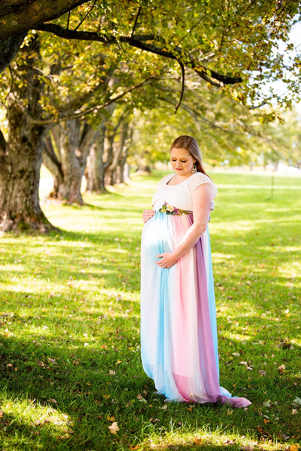 A pregnant woman in a flowing pink dress gently cradles her baby bump in a sunlit park with autumn trees.