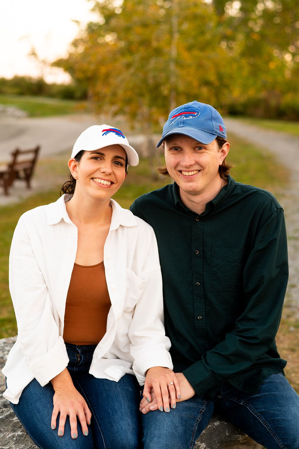 A couple sits together outdoors on an autumn day wearing casual attire and baseball caps.