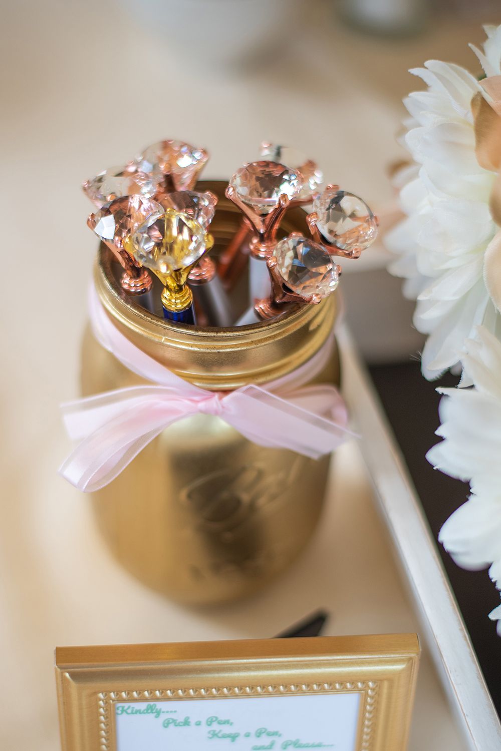 Gold mason jar decorated with pink bow and sparkling rose gold lollipops with white flowers in background.