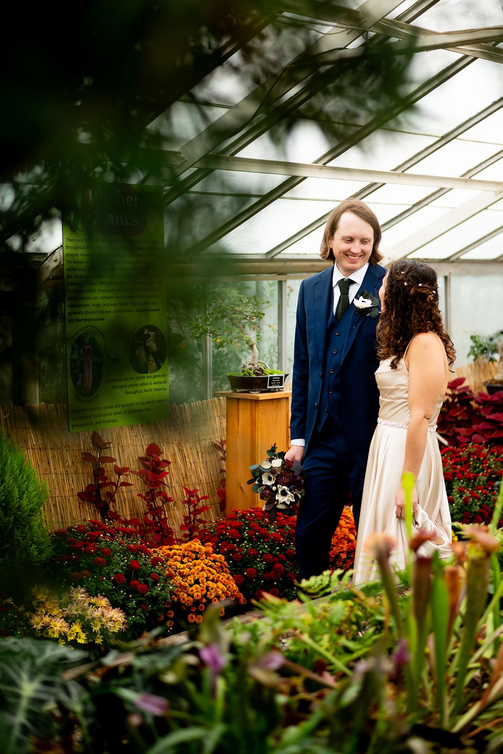 Wedding couple poses in greenhouse filled with vibrant orange and red flowers and greenery.