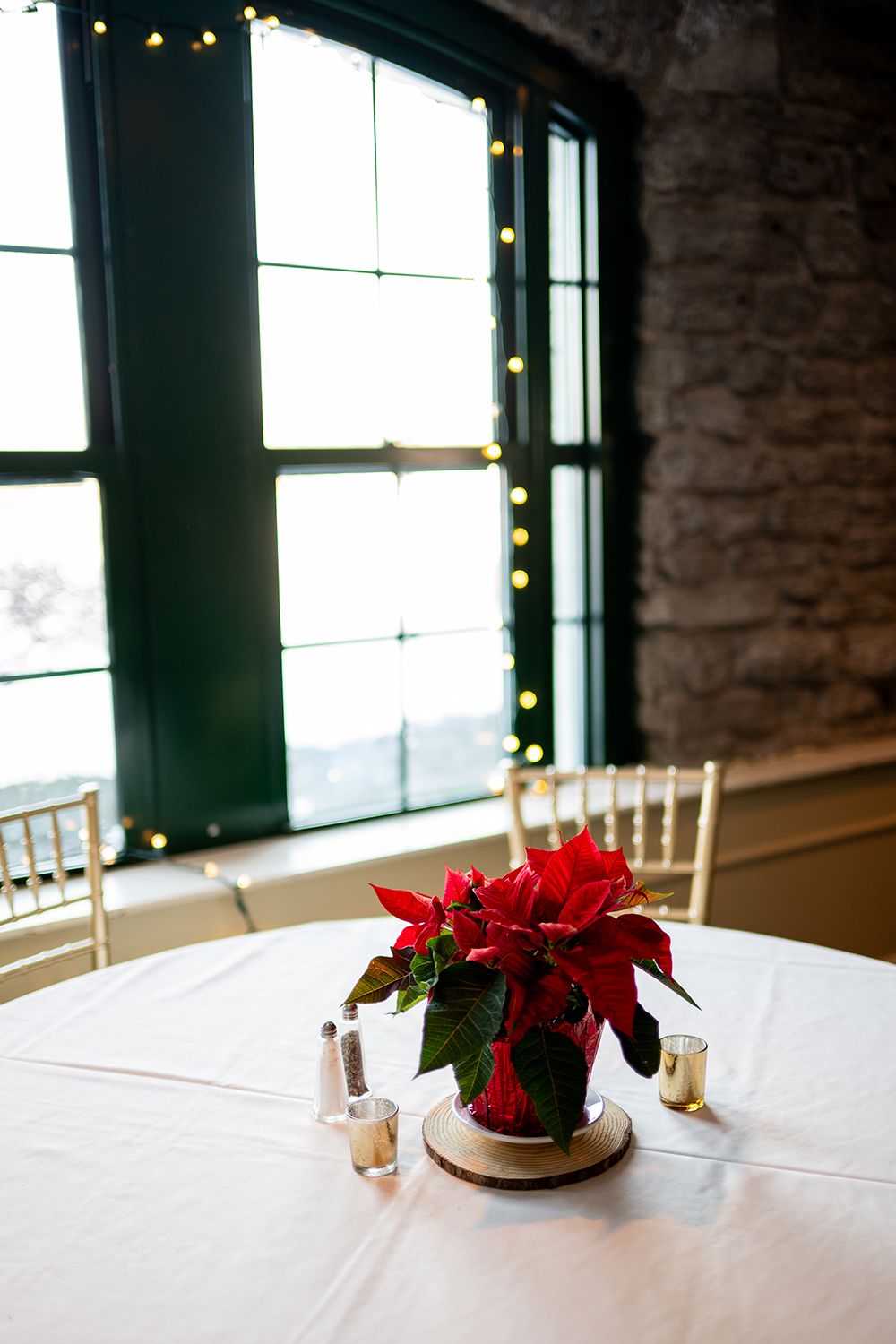 Red poinsettia centerpiece decorations on white banquet tables in an industrial venue with brick walls and holiday lights.