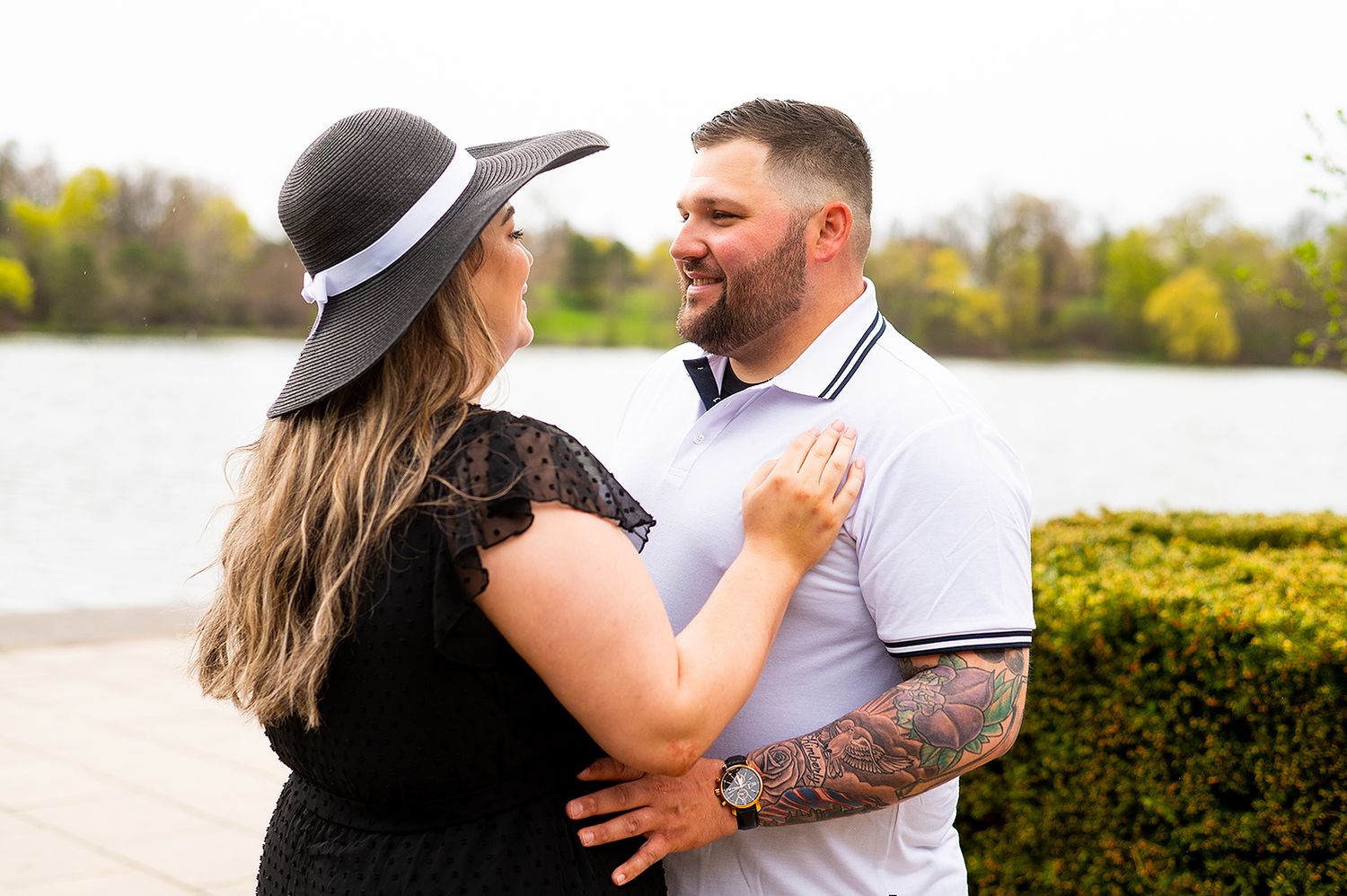 A couple in contrasting black and white outfits share an intimate moment by a lake with sunlit trees behind them.
