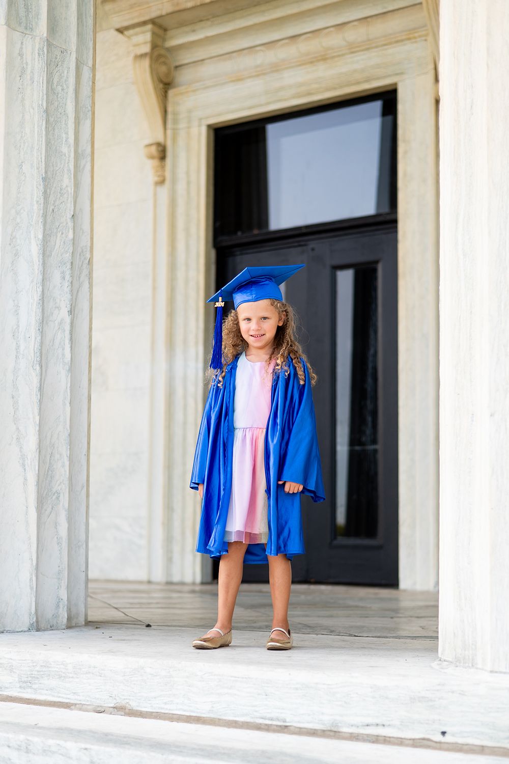 A young graduate in a blue cap and gown stands on a white marble building entrance while wearing pink and sandals.