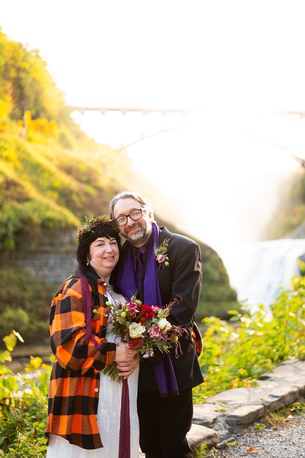 A couple poses together in autumn sunlight by a waterfall, with one wearing a plaid flannel and holding a floral bouquet.
