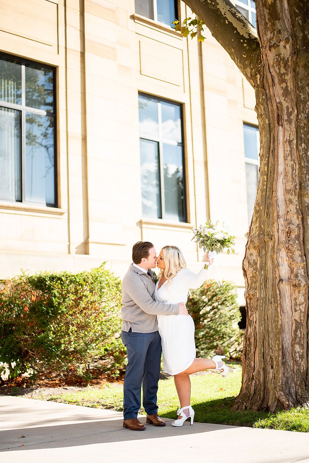 A couple shares a romantic moment beside a large tree in front of a classic stone building on a sunny day.