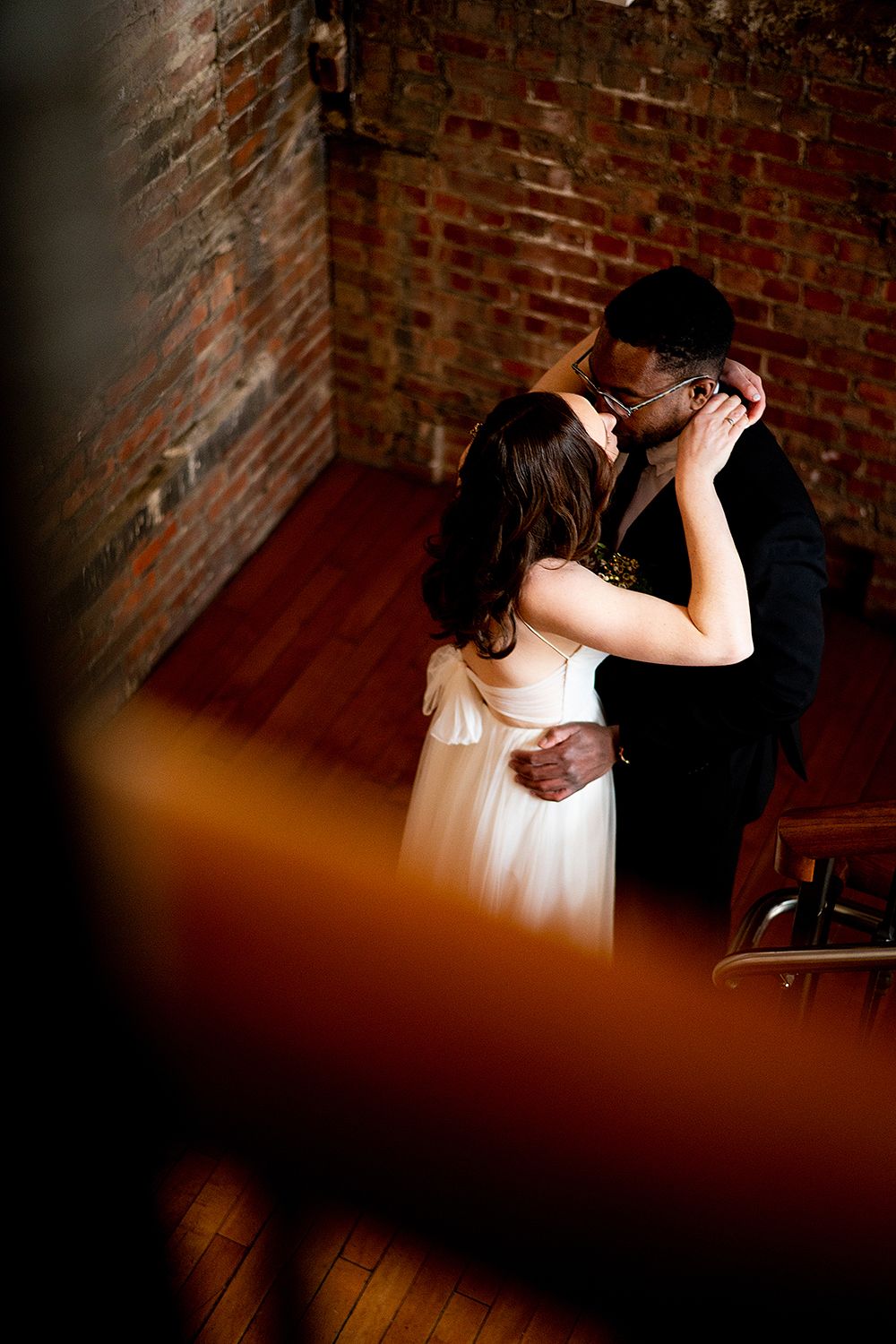 A romantic couple sharing an intimate moment against a rustic brick wall in moody lighting.
