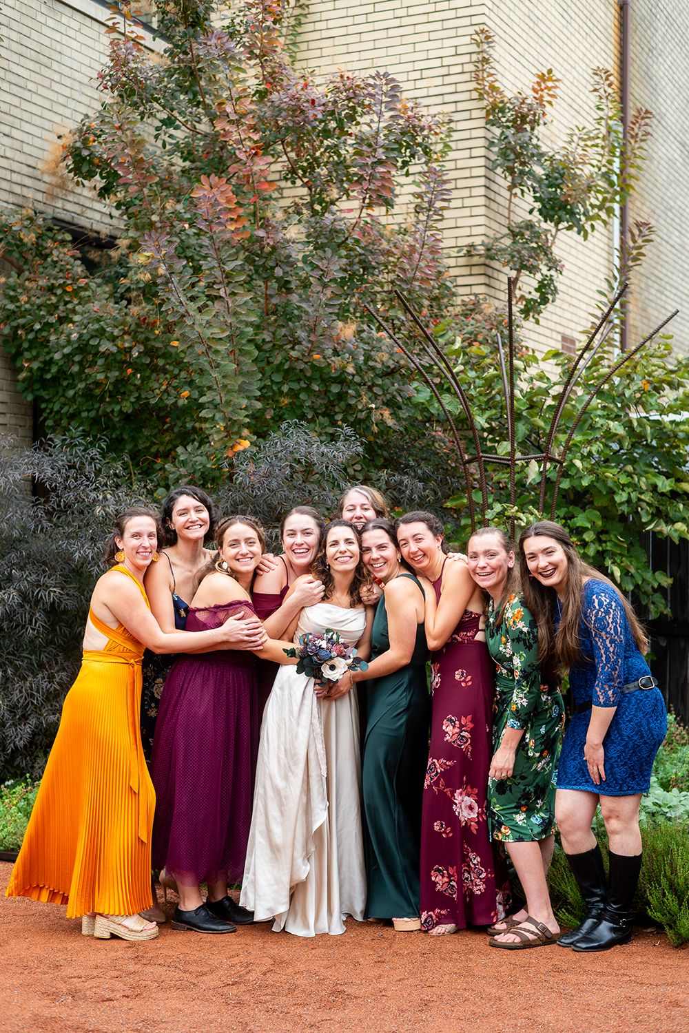 Wedding party group poses in colorful dresses against floral garden backdrop with mix of jewel tones and orange accents.