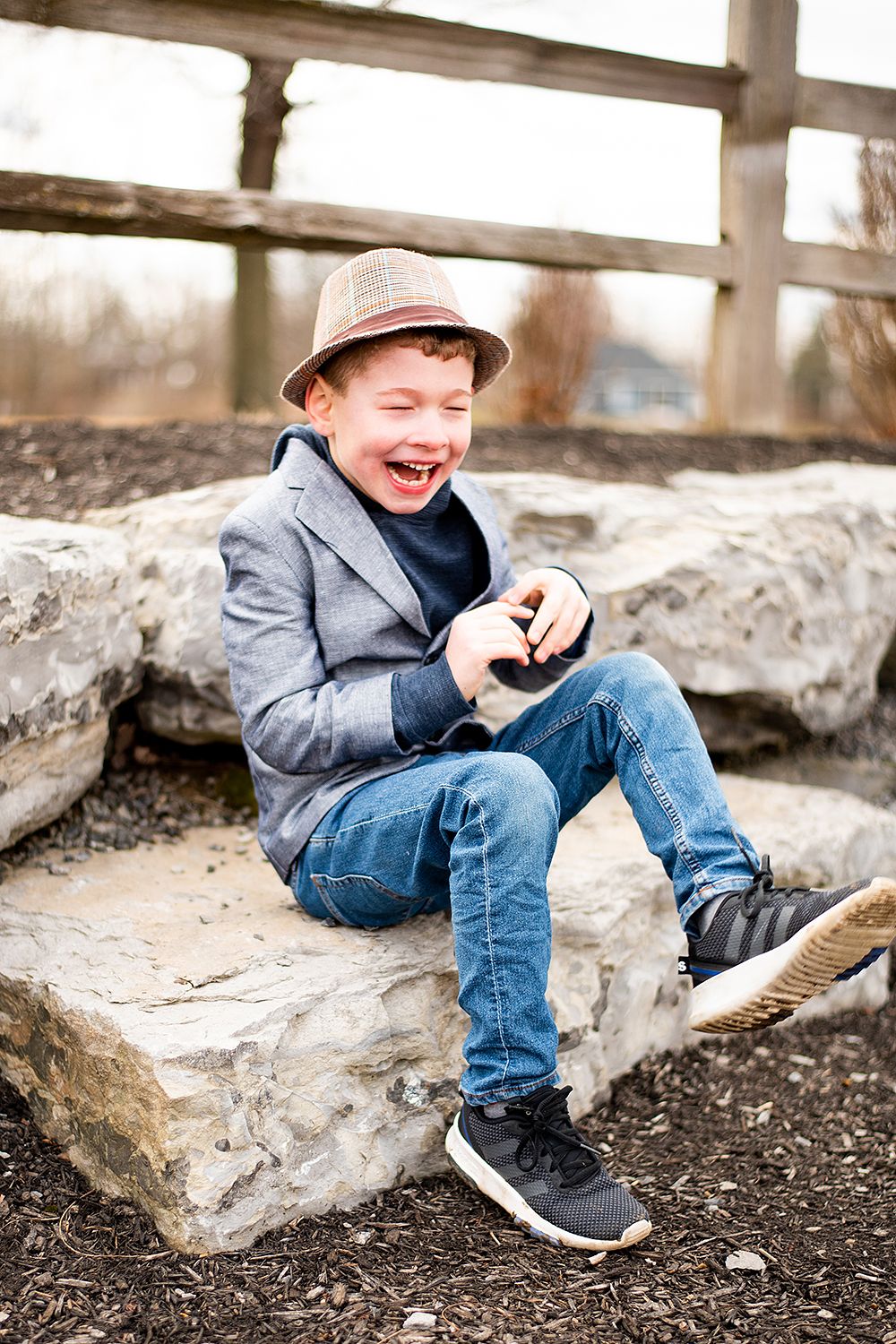 Young child in a gray blazer and fedora hat laughs joyfully while sitting on large rocks outdoors.