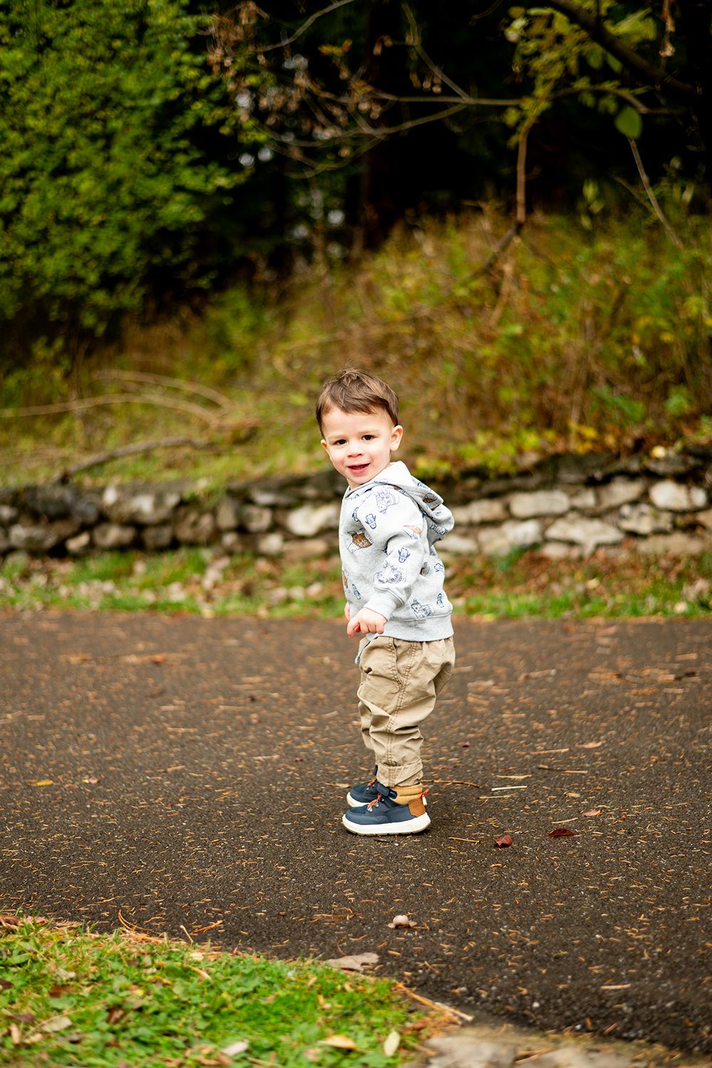 Small child in grey hoodie and khaki pants stands on a dark pathway near a stone wall and greenery.