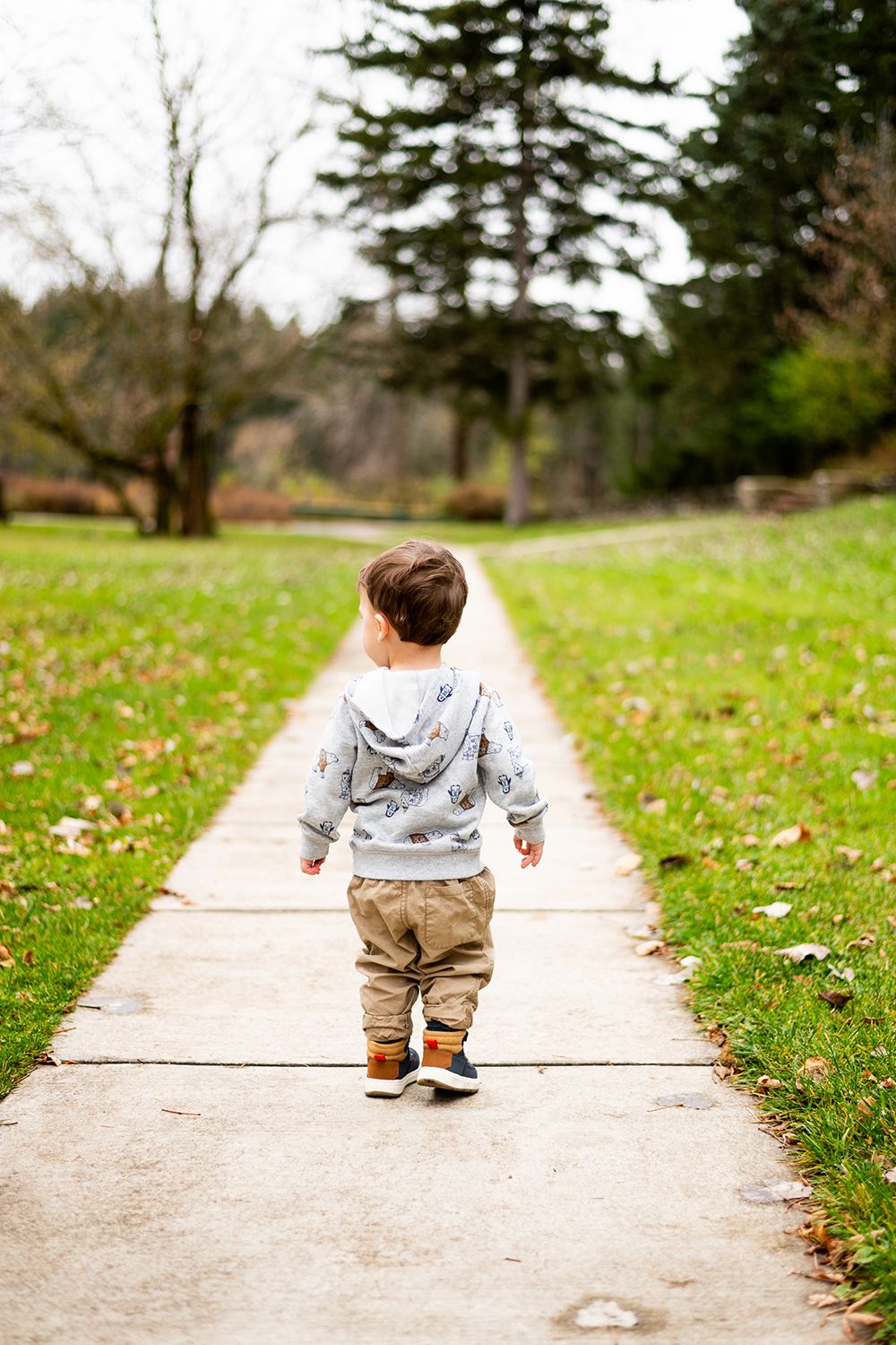 Small child in gray hoodie and khaki pants walking alone on concrete path through grassy park area.