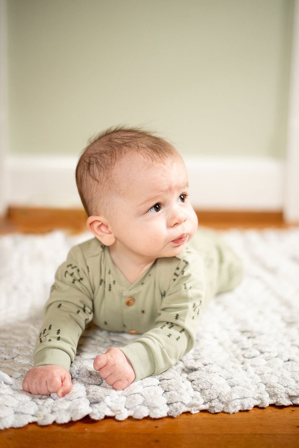 A baby wearing a sage green patterned onesie lies on a white textured rug on hardwood flooring.