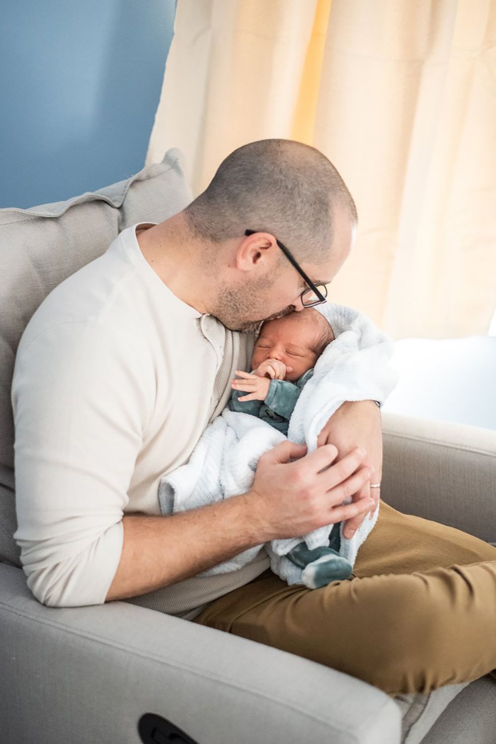 A tender moment shows a parent in glasses and white sweater cradling a newborn baby wrapped in blankets on a gray couch.