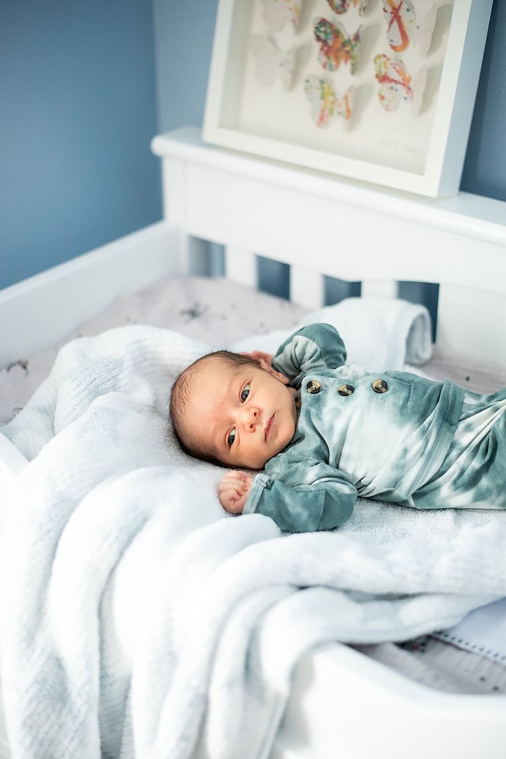 Baby in soft blue sleeper rests on white bedding in a nursery decorated with framed art on the wall.