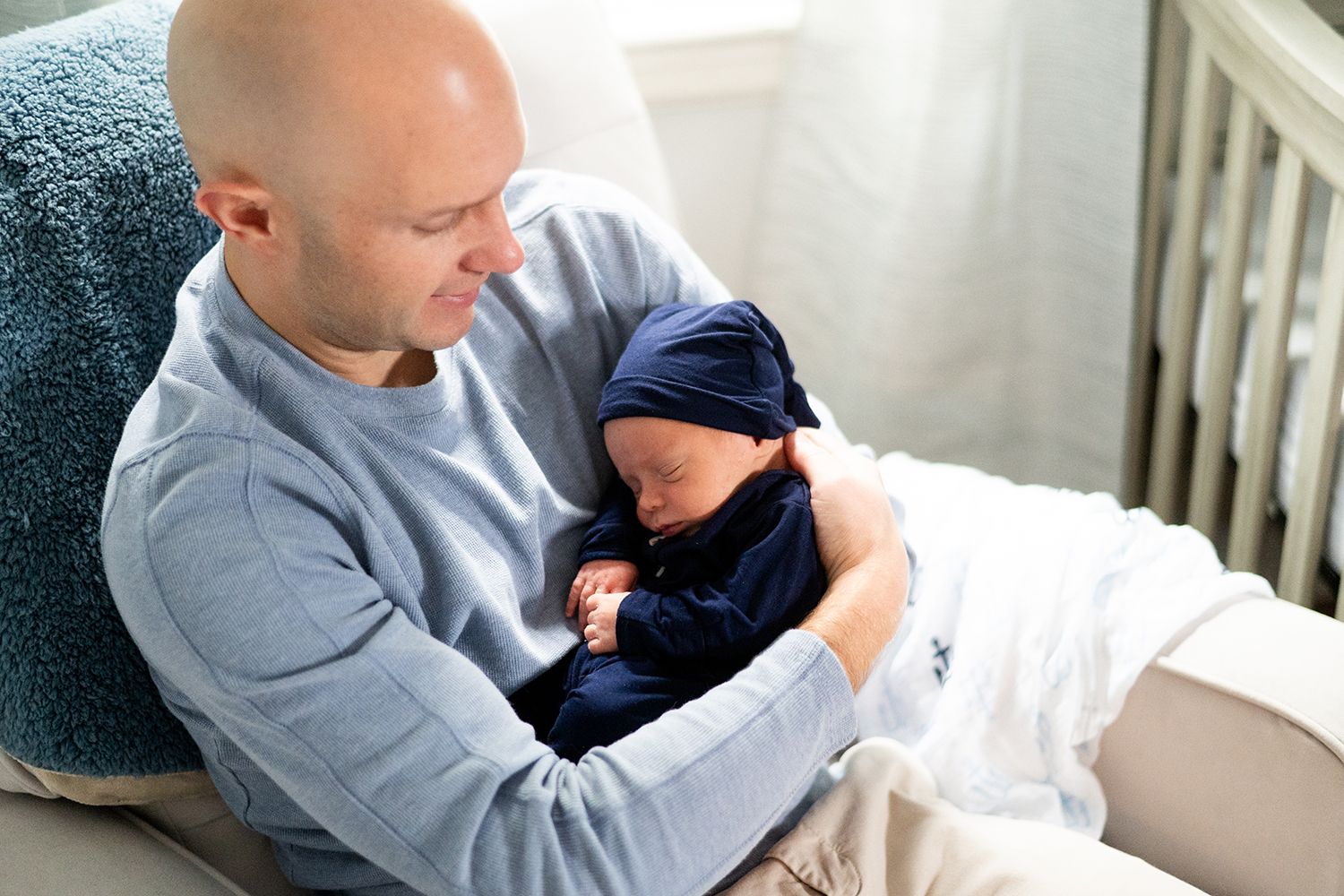 Tender moment as parent holds sleeping newborn baby wearing navy outfit while sitting on couch in natural light.