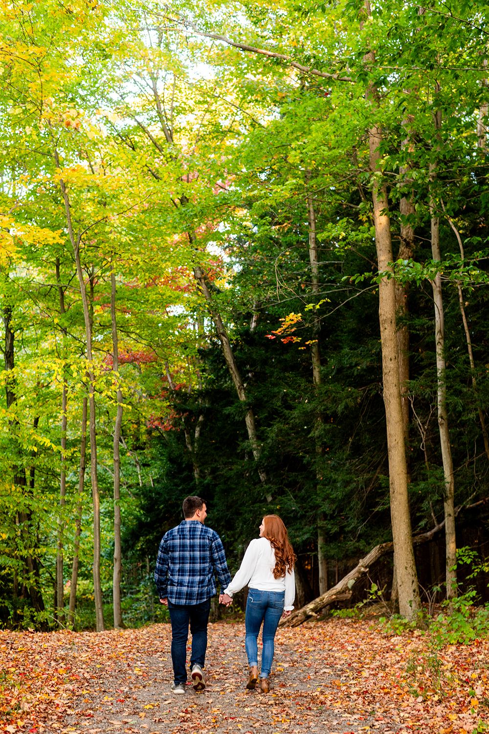A couple walks along a scenic autumn trail surrounded by tall trees with golden yellow leaves falling on the ground.
