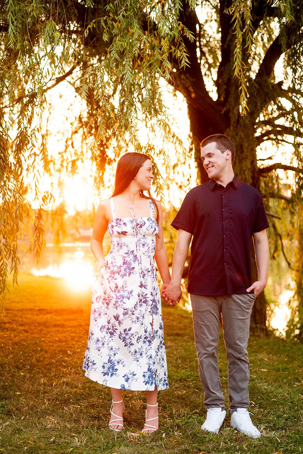 A couple stands together under a willow tree during golden hour sunset with warm glowing light filtering through branches.