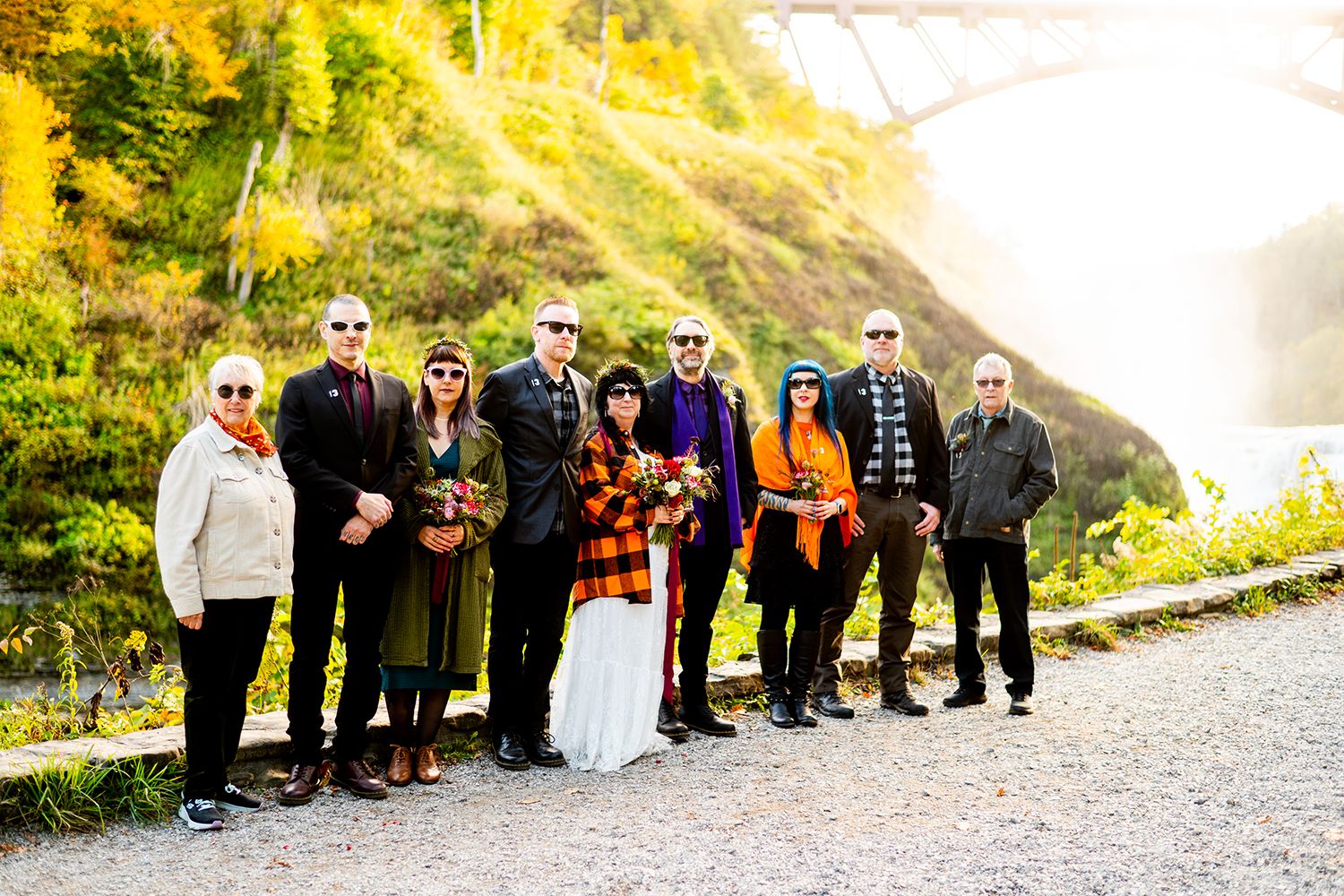 A group of people in warm winter clothing stand together on a path with autumn foliage and mountains in the background.