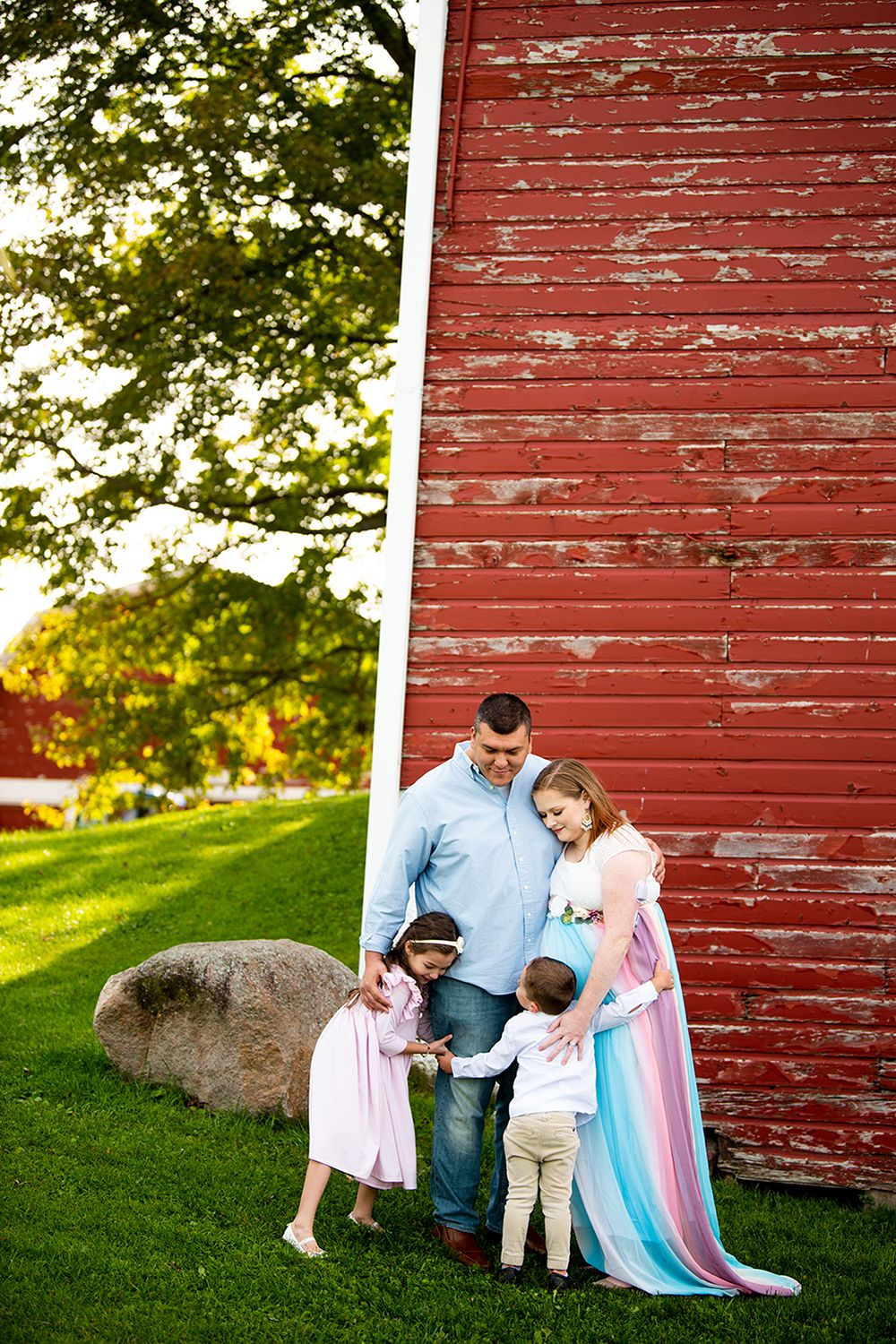 Family stands beside a rustic red barn wall with large boulder and green grass during golden hour.