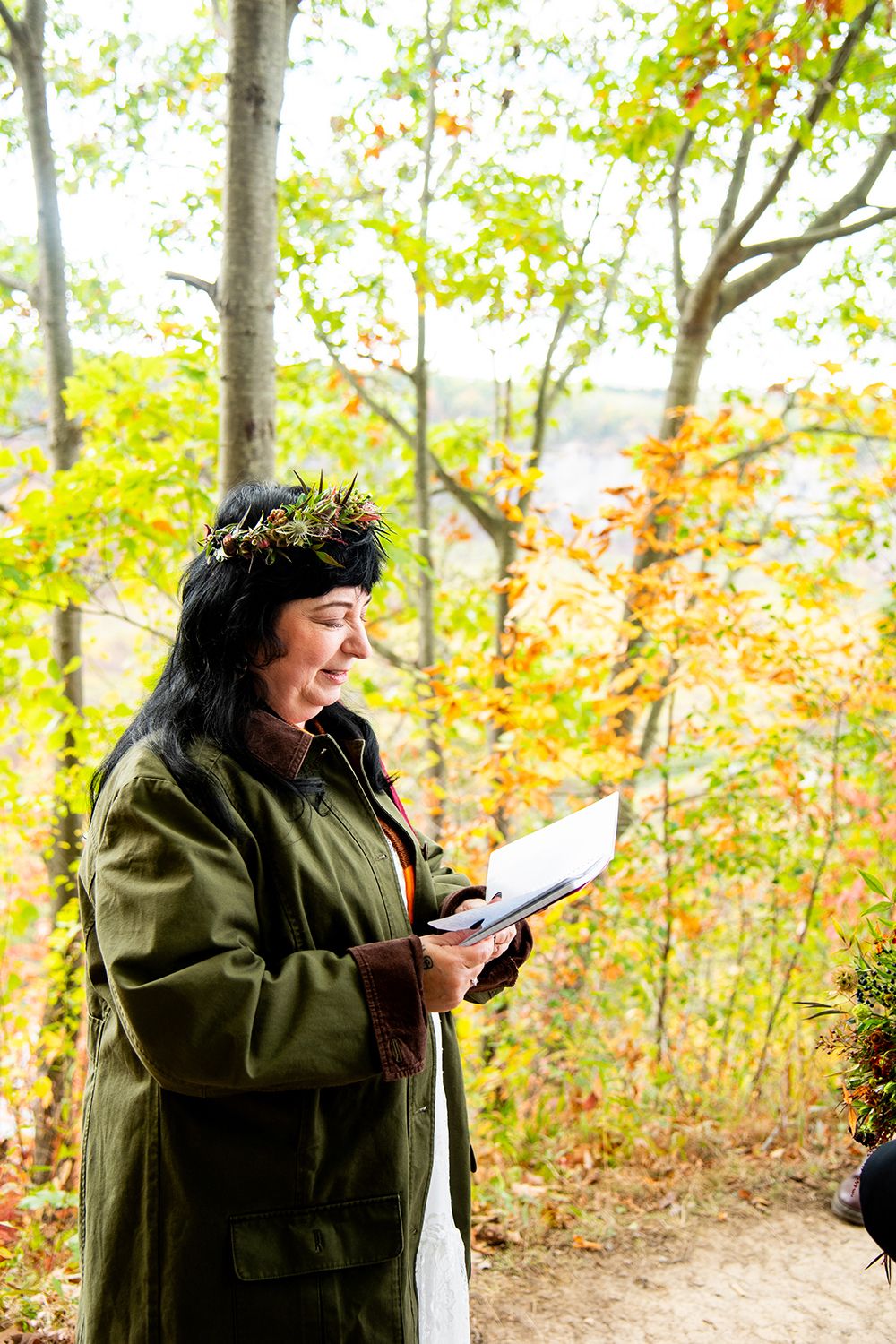 Person in a green coat and nature-inspired crown reads from a book in an autumn forest setting.