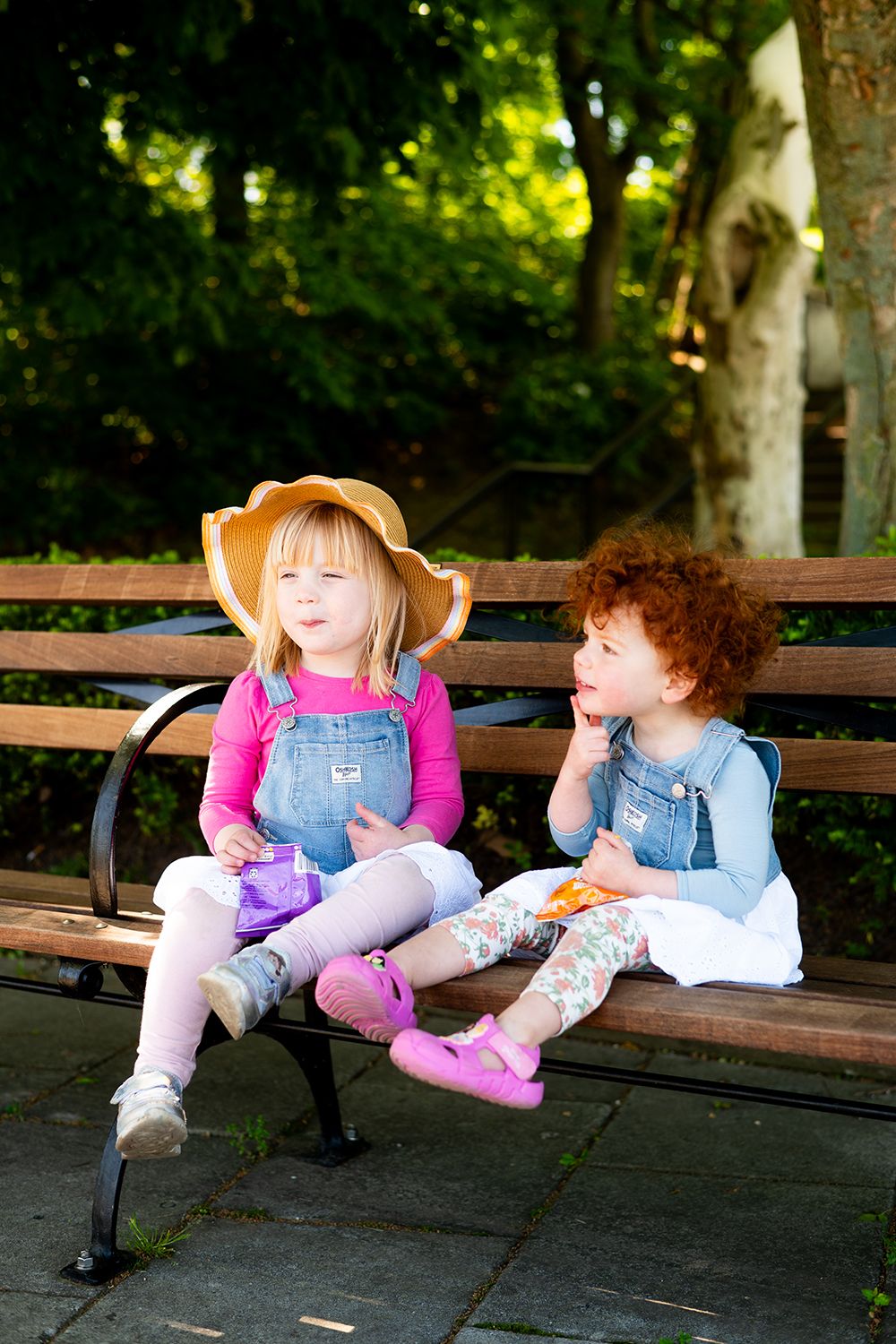Two toddlers in overalls and sun hats sitting together on a park bench having a conversation.