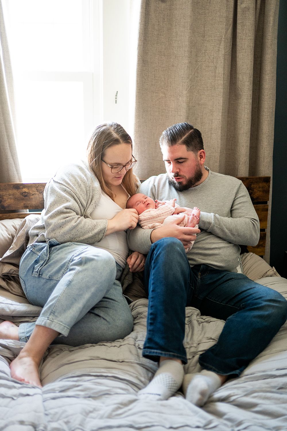 Parents sitting together on bed holding and looking lovingly at their newborn baby.