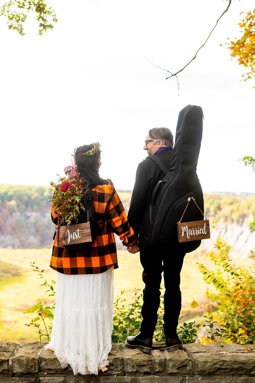 Couple sitting on stone wall in autumn with orange plaid flannel shirt and white dress during outdoor photoshoot.