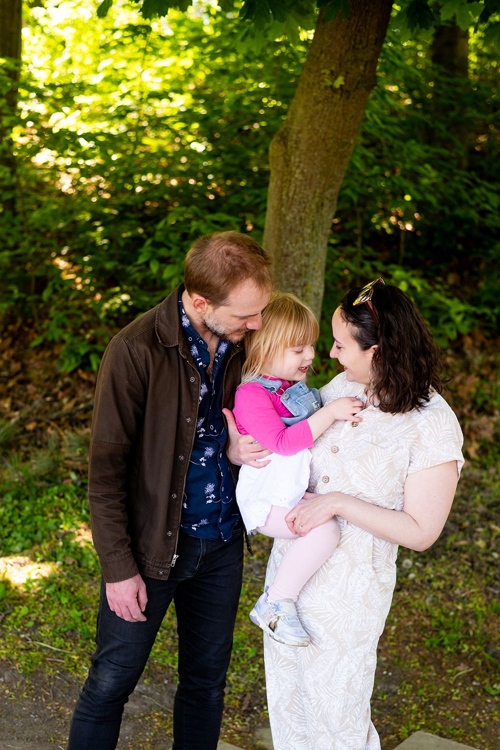 A couple shares a tender moment with their small child in a lush green forest setting on a sunny day.