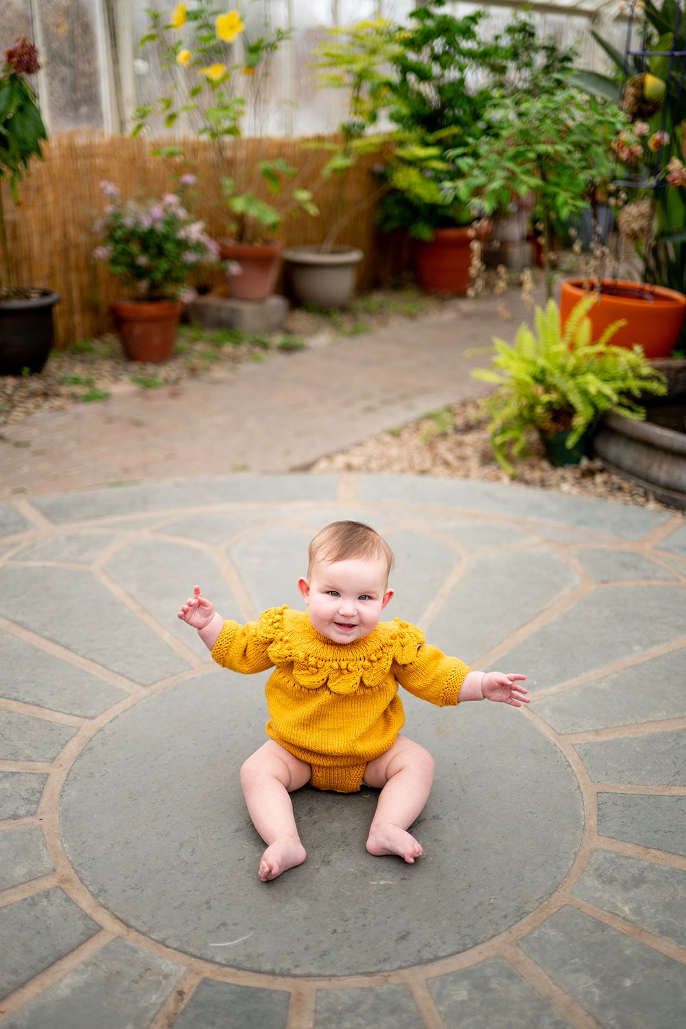 A baby in a mustard yellow ruffled outfit sits and plays on a circular stone patio pattern.