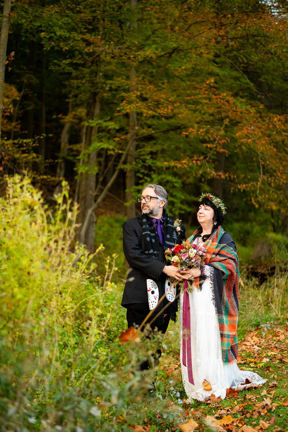 A couple in traditional wedding attire share an intimate moment in a wooded setting with autumn foliage.