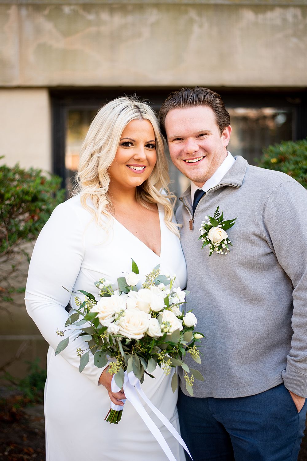 A couple poses together outdoors for a wedding photo, with the bride holding a white and green floral bouquet.
