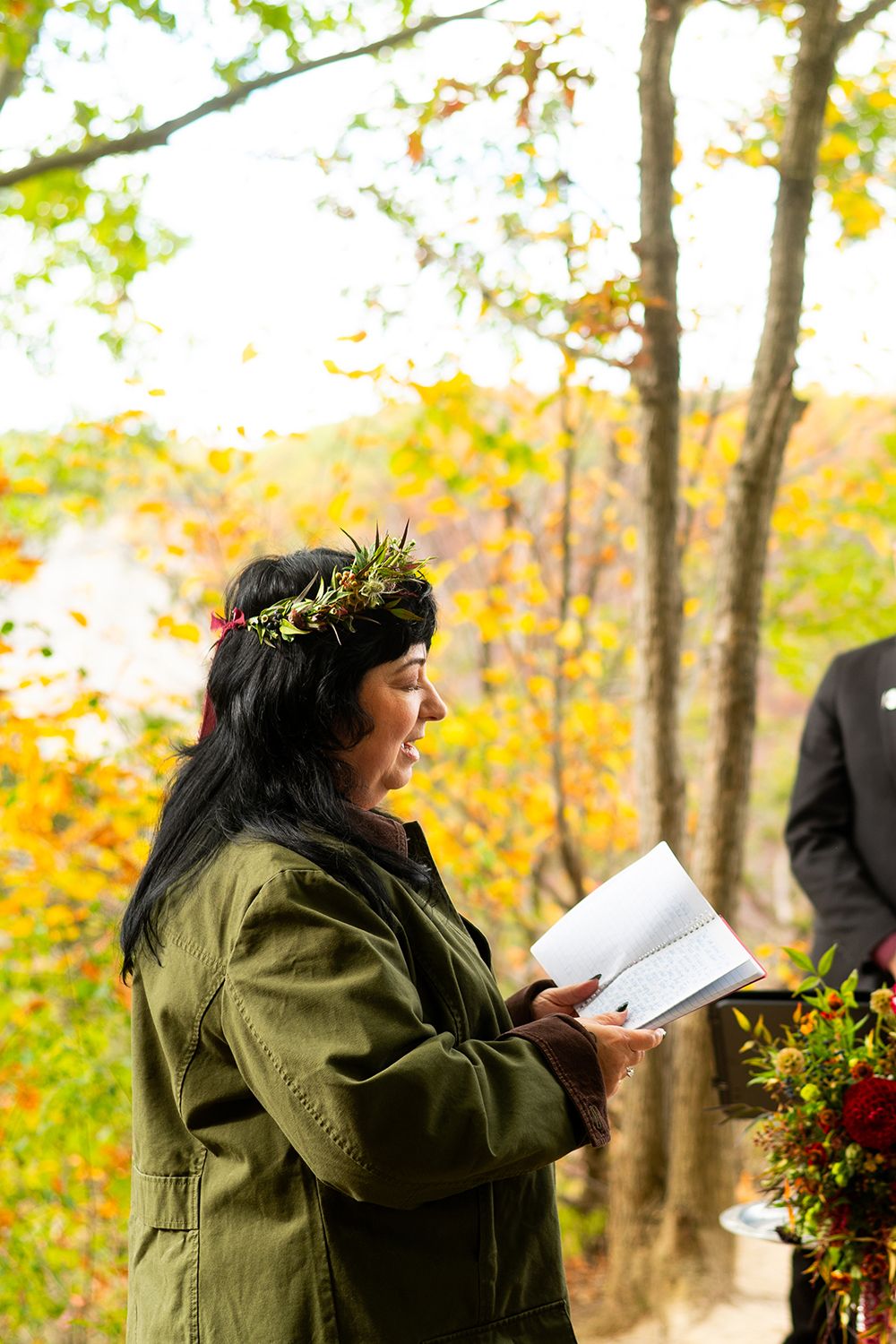 Person in olive green coat stands in autumn woods while holding papers and colorful bouquet of flowers.