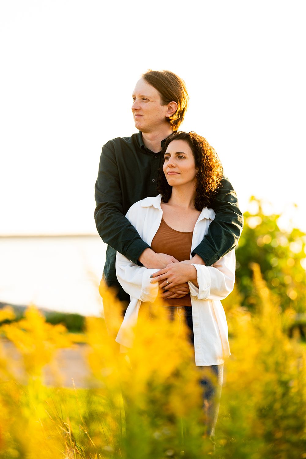 A couple embraces in a golden sunlit field during sunset, surrounded by yellow wildflowers.