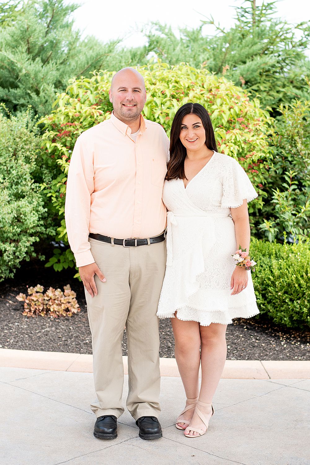 Two people standing together outdoors in light-colored outfits with green foliage in the background.