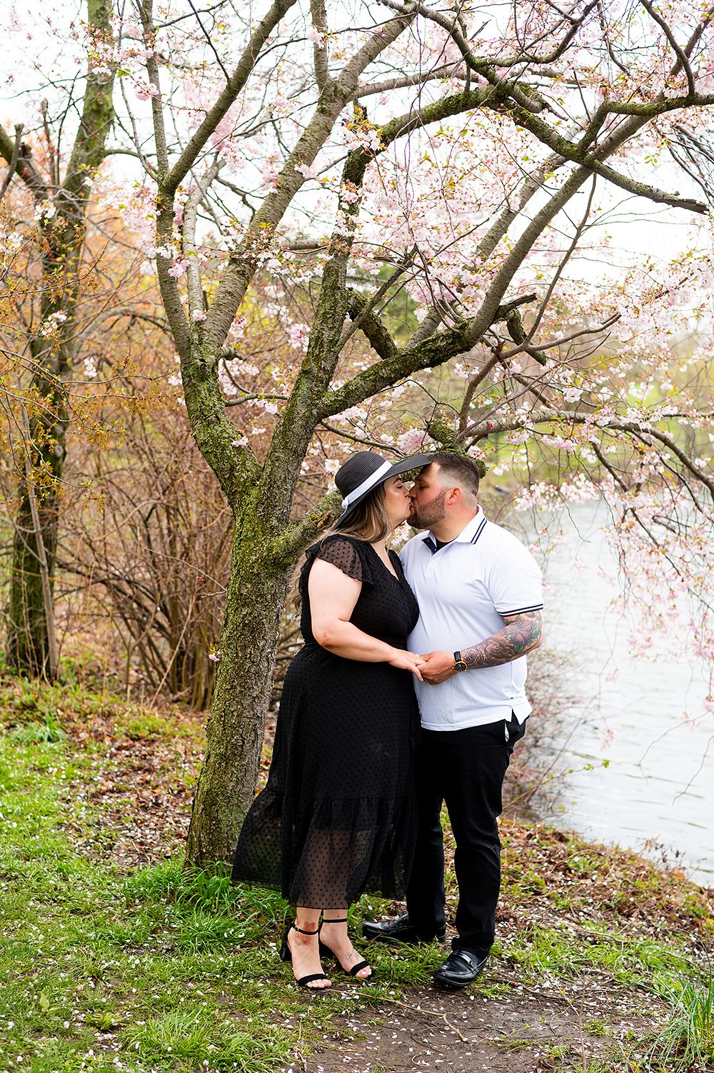 A romantic couple shares an intimate moment under blooming cherry blossom trees beside a serene lake.
