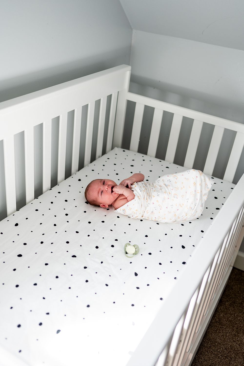 A white crib with polka dot bedding sits in a room with dark carpeting as seen from multiple angles.