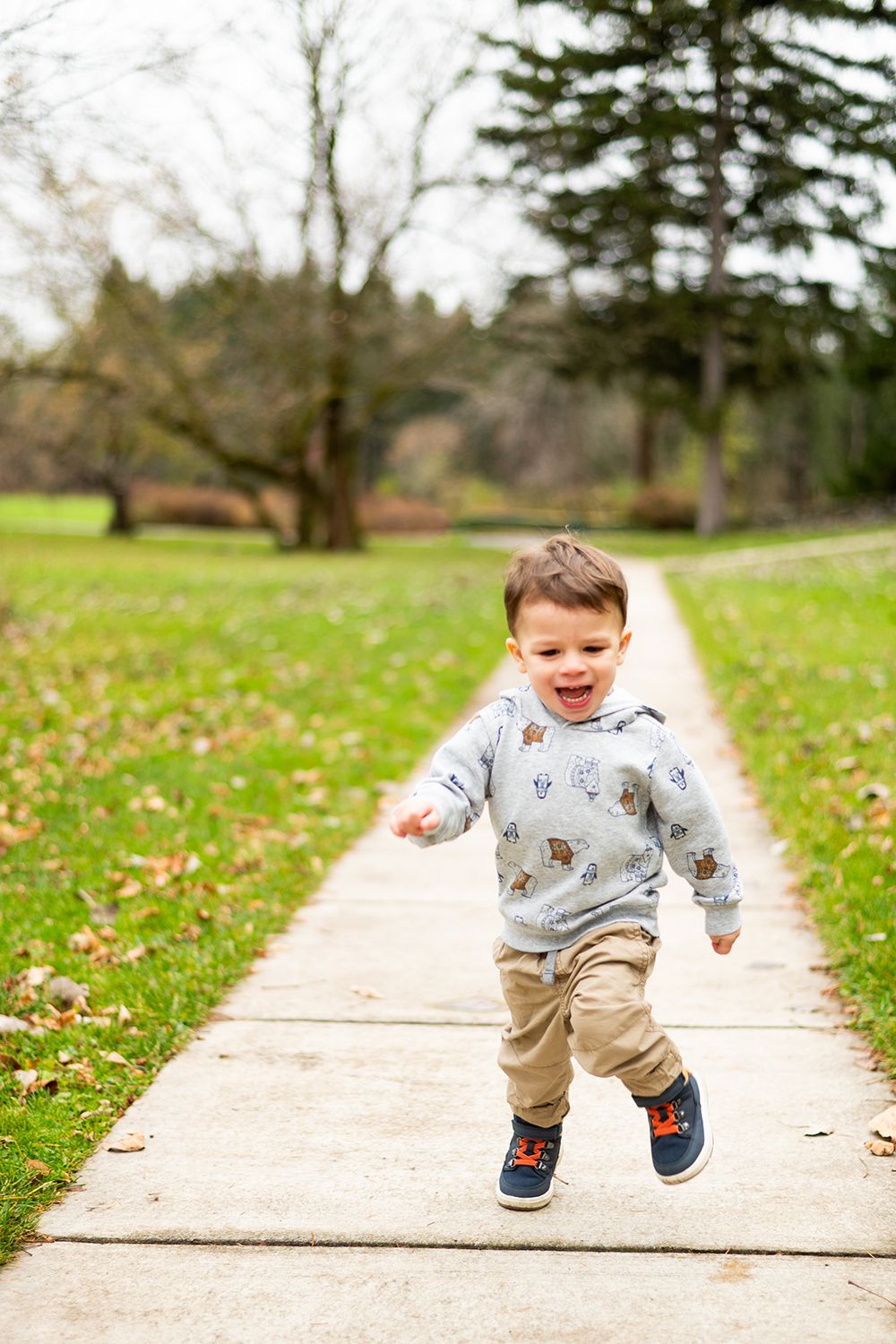 Young child in a patterned sweater and khaki pants joyfully running on a concrete path surrounded by grass and fallen leaves.