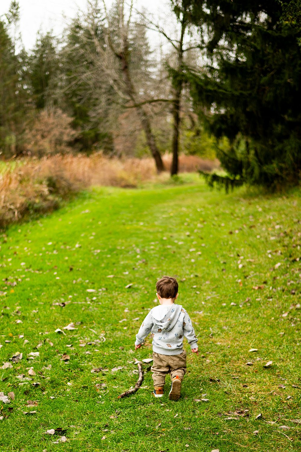 Child running on grass path through wooded area wearing gray hoodie and tan pants.