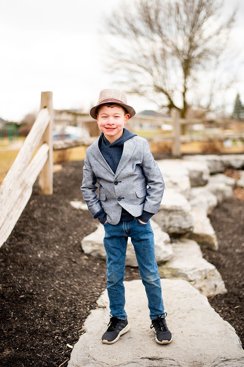 Someone wearing a gray blazer, pink hat and jeans stands confidently on a stone path in an outdoor garden setting.