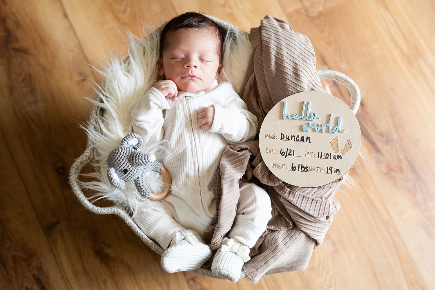 Newborn baby rests peacefully in white sleeper on fur blanket next to milestone marker and stuffed animal.