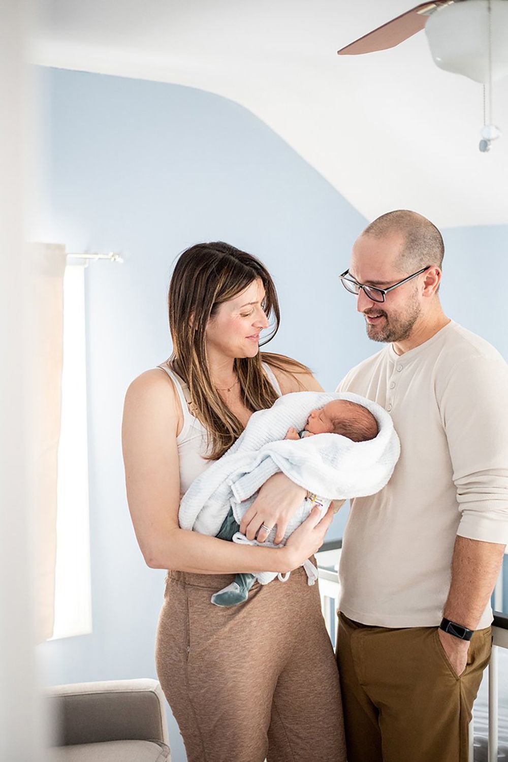 A couple stands together in a bright room while holding a newborn baby wrapped in a white blanket.