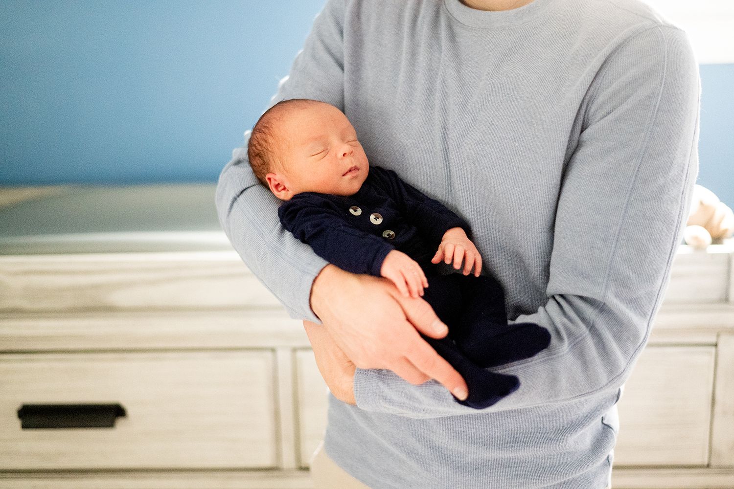 Newborn in navy outfit resting peacefully against gray sweater in sunlit nursery.