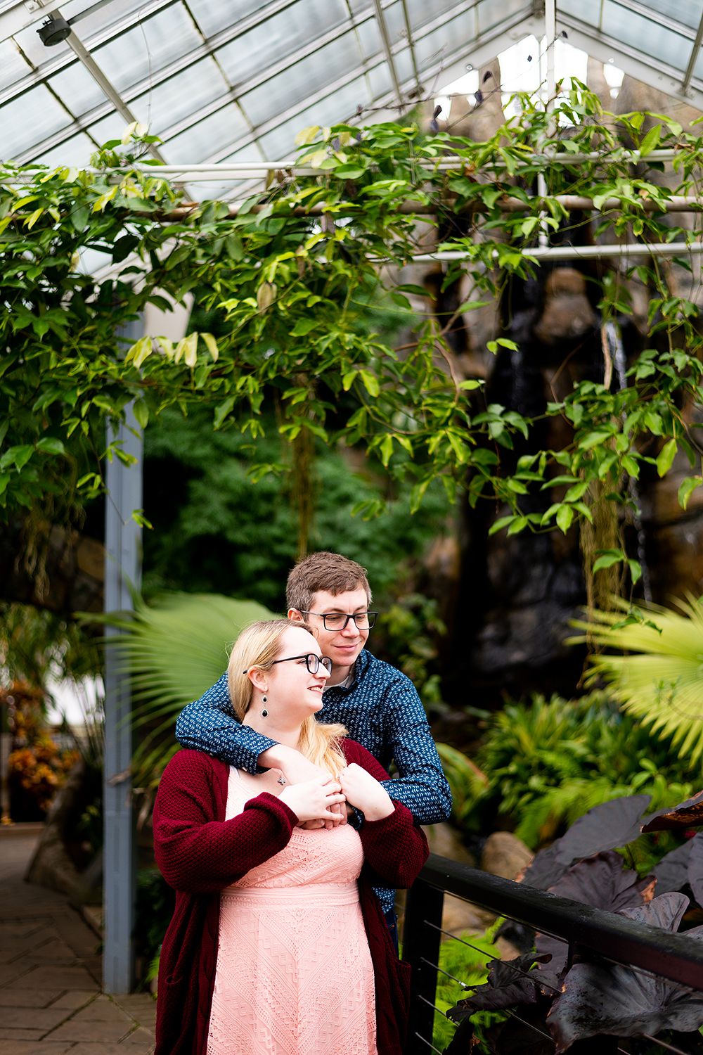 A loving couple shares a romantic moment in a sunny greenhouse filled with lush greenery and tropical plants.