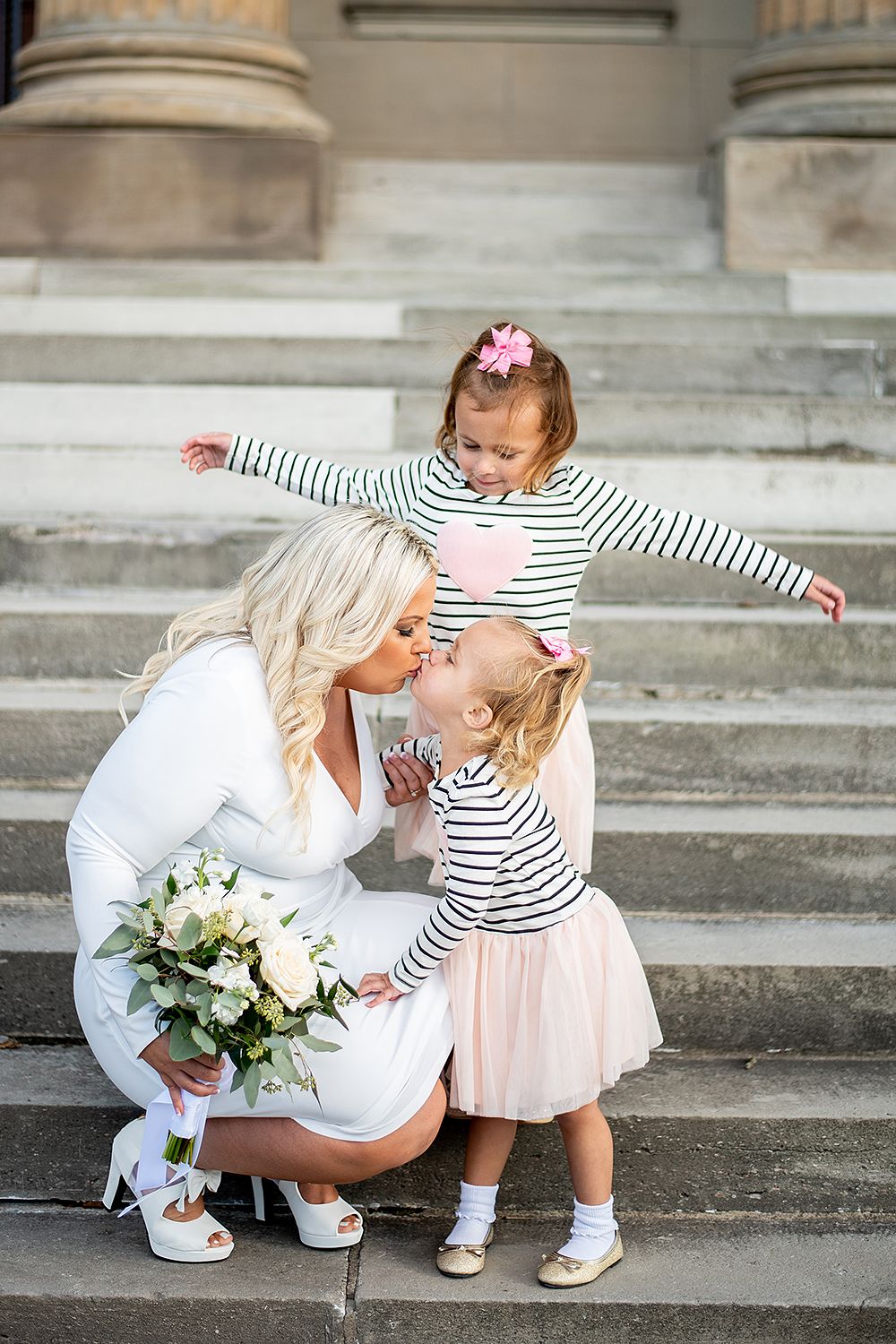 Mother in white dress holding bouquet embraces children wearing matching striped shirts on stone steps outdoors.