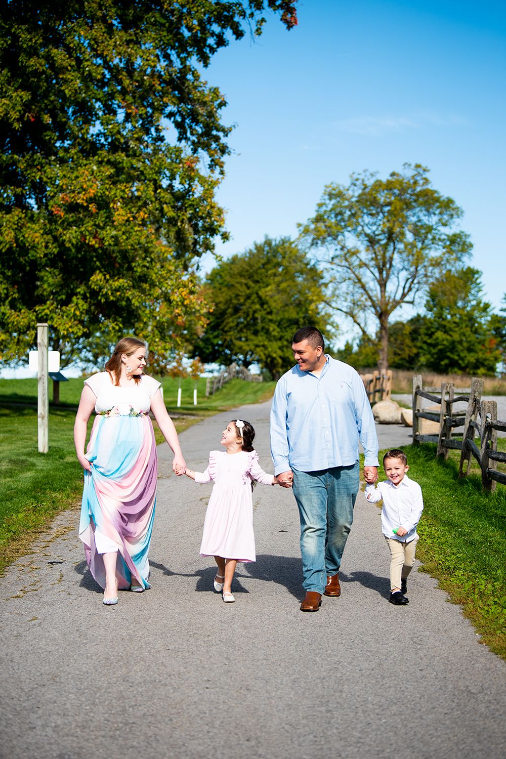 A family takes a stroll together on a sunny day along a tree-lined country path with white fencing.